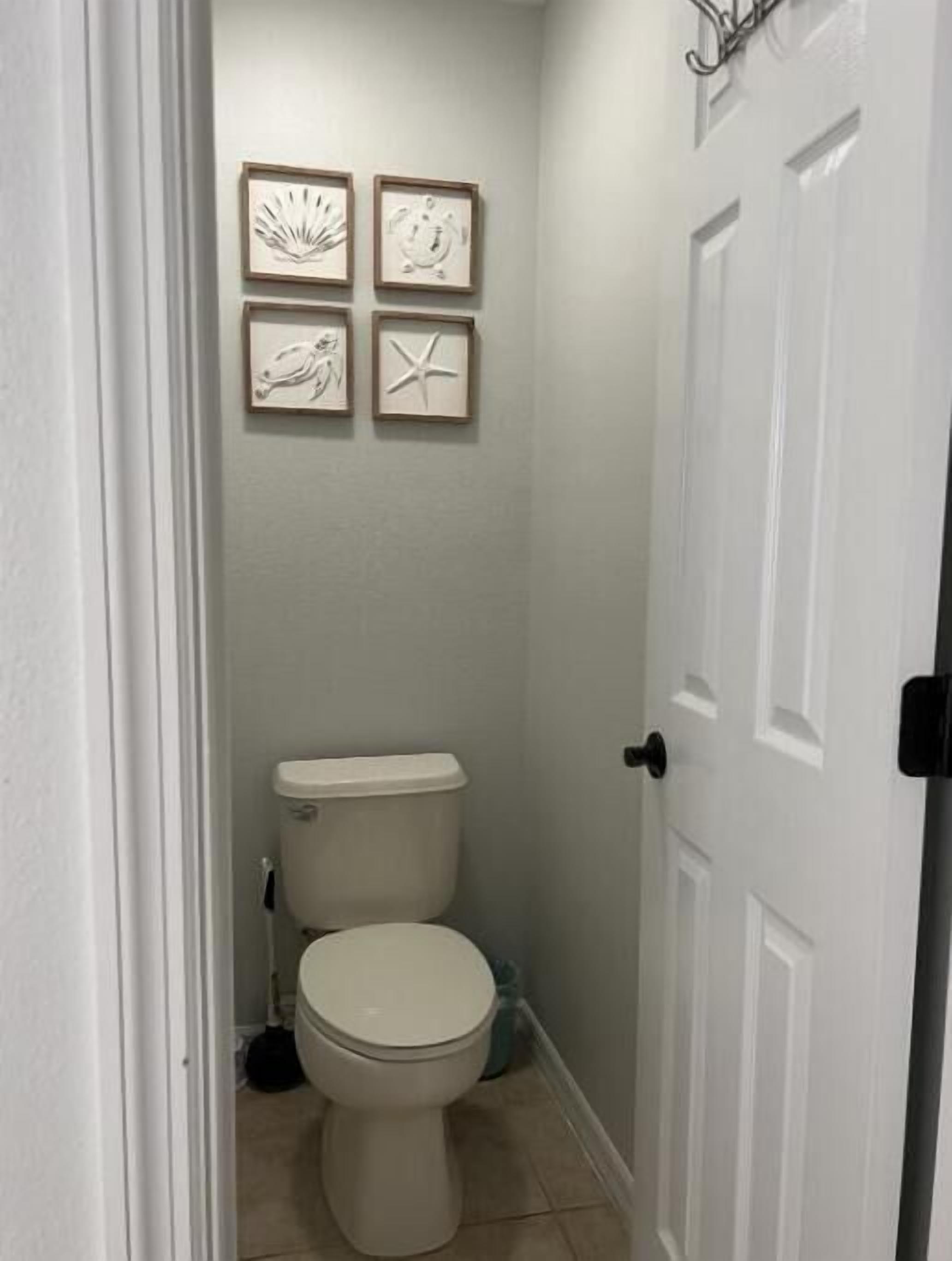 A small, clean bathroom with a toilet, a broom, and a trash can. There are four framed pictures on the wall.
