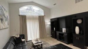 A modern living room with a black leather sofa, a ceiling fan, and a large window with curtains. - Luxury 5bd home close to Disney parks private gated property  (Kissimmee)