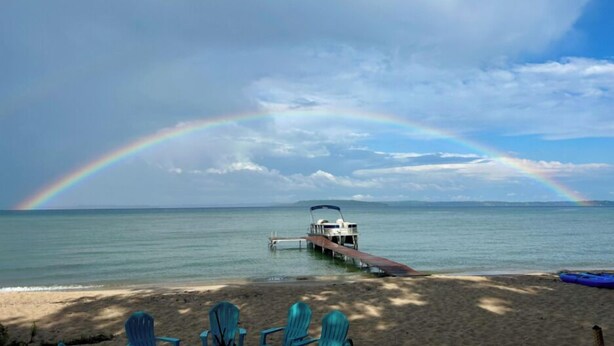 Een strand met een boot afgemeerd aan de steiger, een regenboog aan de hemel en turquoise stoelen in het zand.