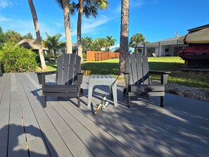 A deck with two Adirondack chairs and a small table, surrounded by palm trees and a grassy area.