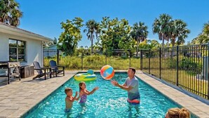 Uma piscina sob um céu ensolarado, com palmeiras e uma pessoa relaxando em uma espreguiçadeira.