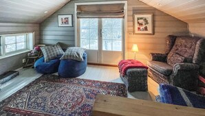 Un acogedor dormitorio en el ático con un ventanal grande, una alfombra estampada, un sillón de peluche azul, un sillón de cuero y una lámpara.