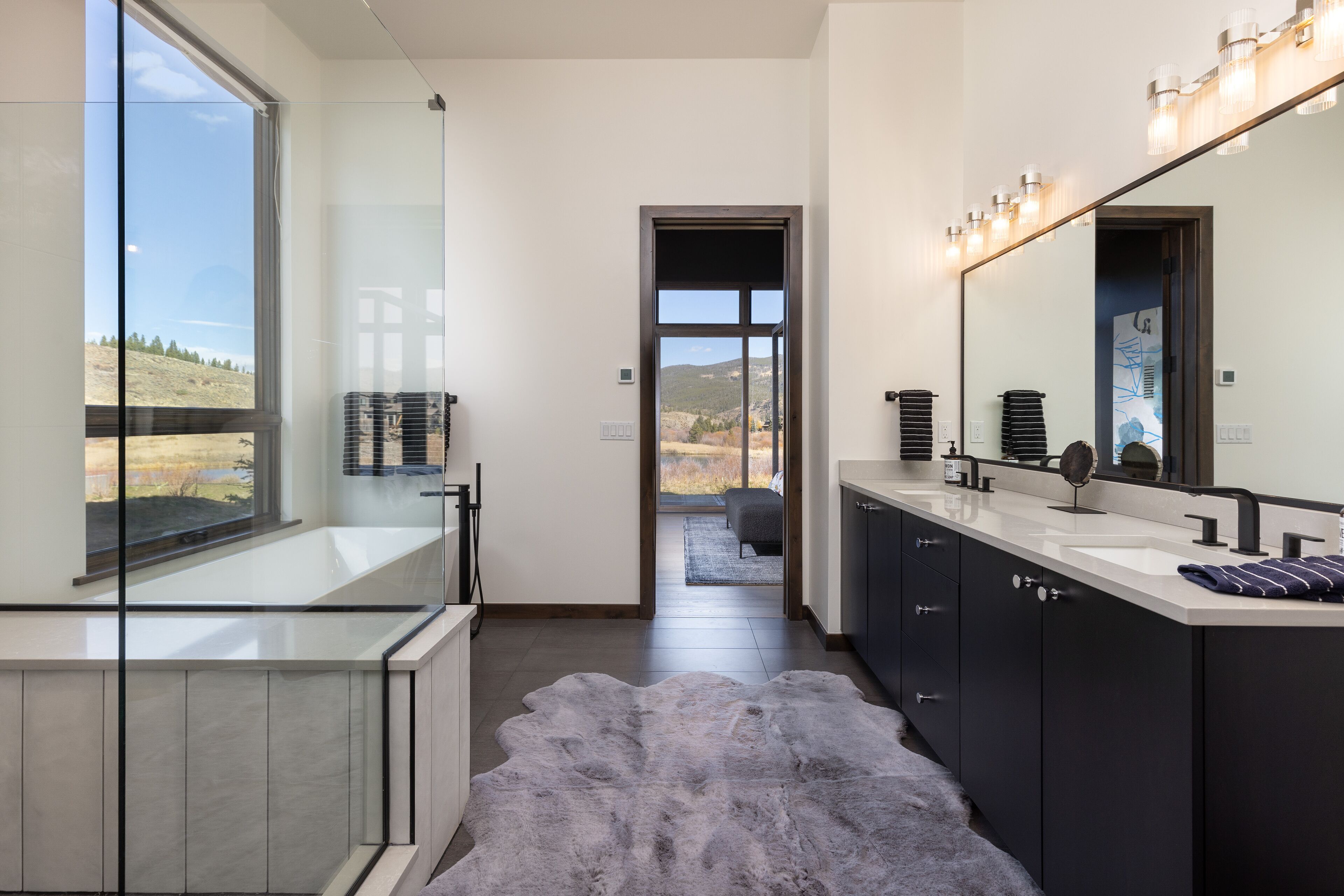Un baño moderno con un ventanal grande, cabina de ducha de vidrio, tocador con espejo y vistas al exterior.