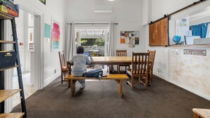 A child sitting at a wooden table in a room with a carpeted floor, a white wall with a clock, and a door leading outside. - Worldwide Backpackers Limited (Wellington)