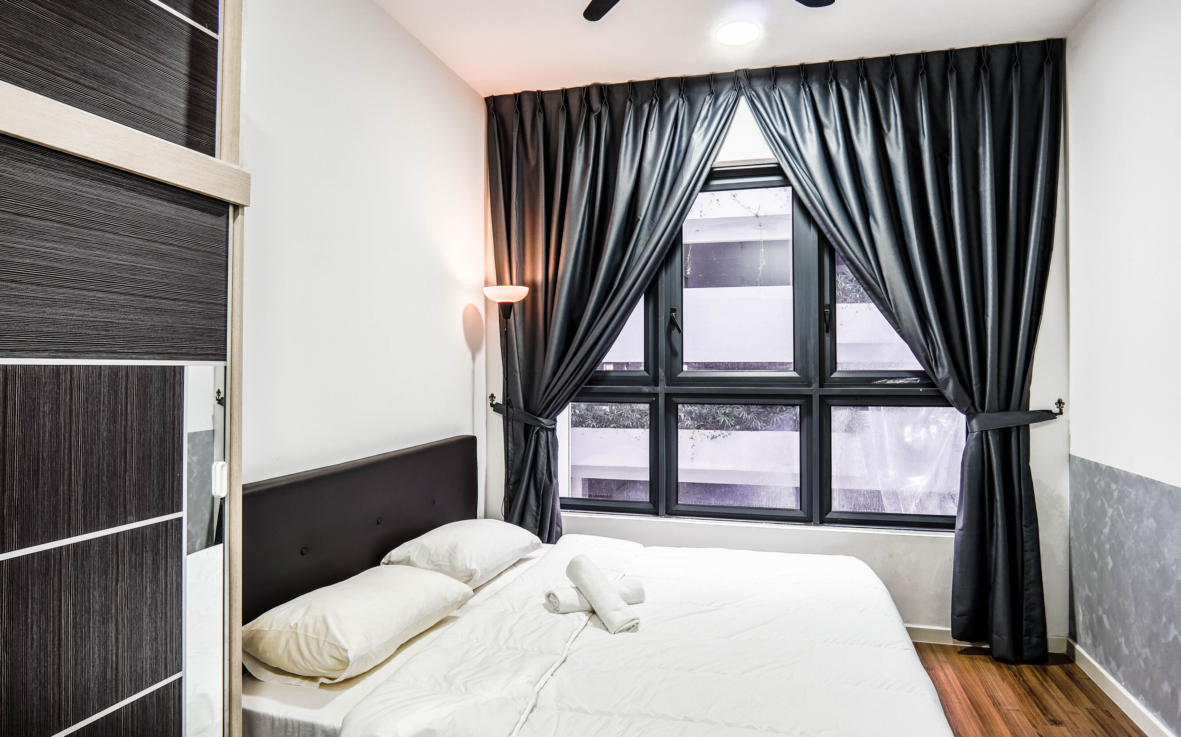 A neatly made bed with white linens and two pillows, a window with dark curtains, and a wall with a dark wood panel design.
