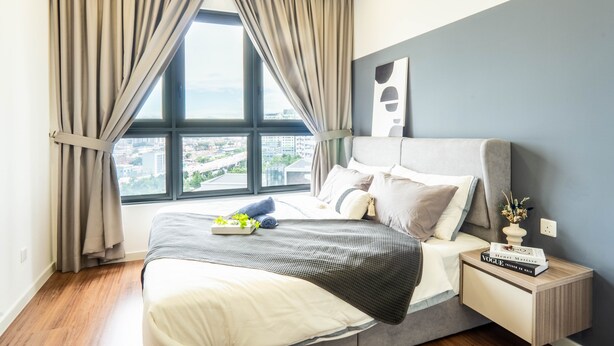 A modern bedroom with a large window, a bed with a grey headboard, and a bedside table with books.