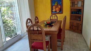 Un coin repas avec une table et des chaises en bois, un tableau représentant des fruits accroché au mur et un meuble contenant des ustensiles de cuisine.