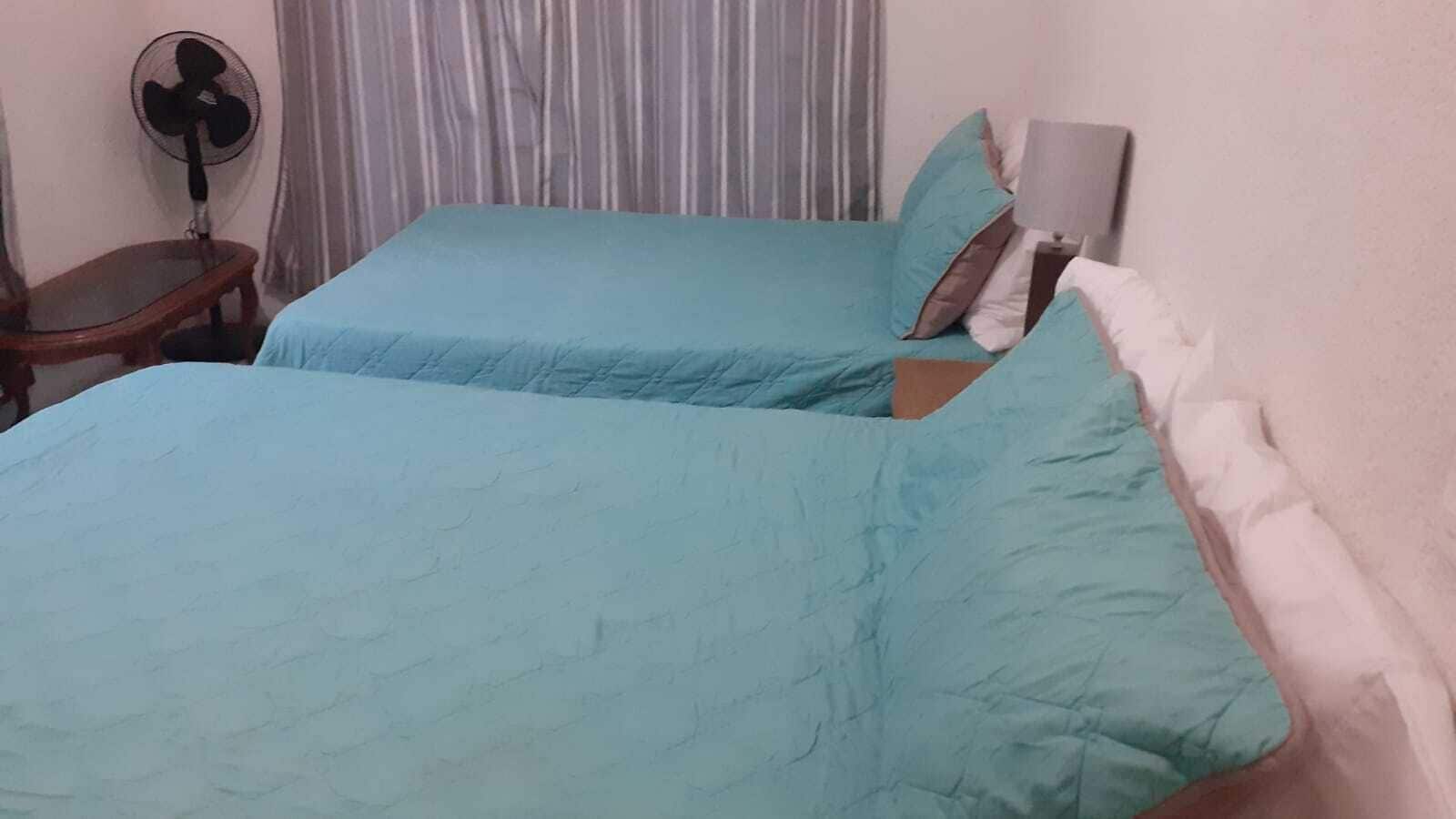 Una habitación con dos camas, una mesita de noche, un ventilador y una ventana con cortinas.