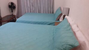 Une chambre avec deux lits, une table de chevet, un ventilateur et une fenêtre avec des rideaux.
