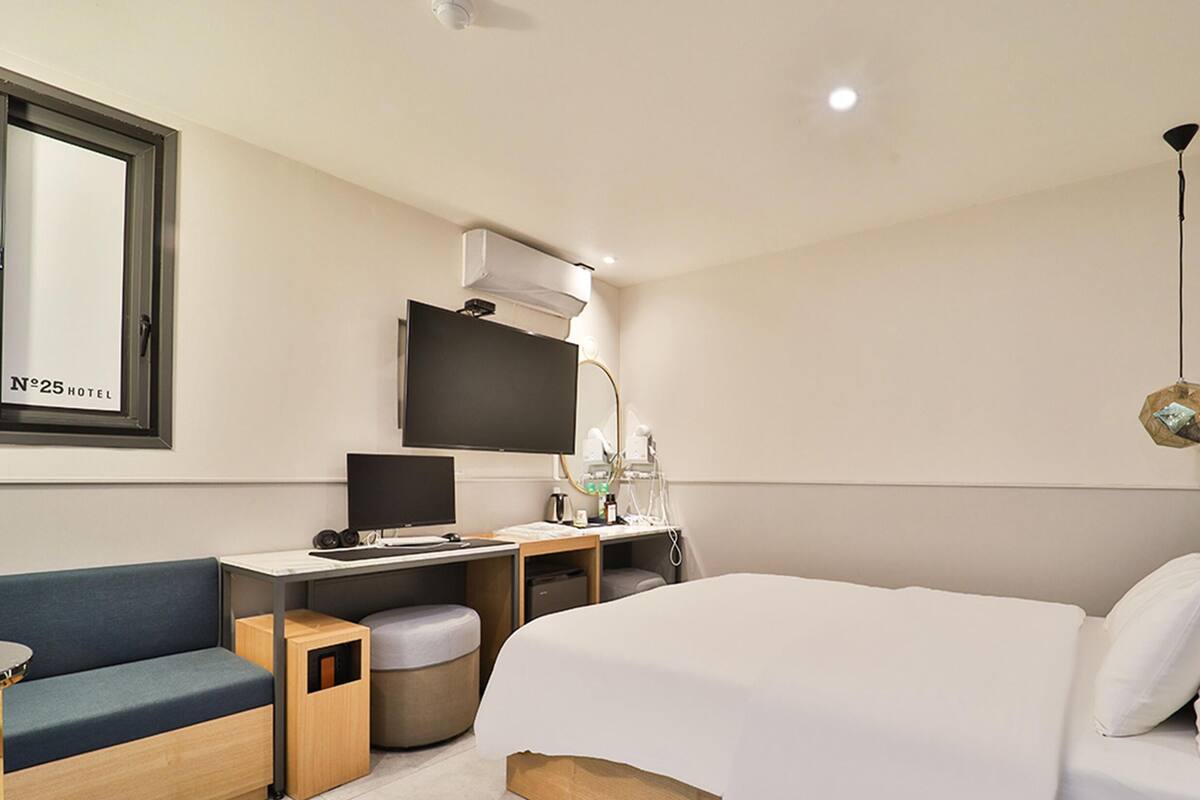 Habitación de hotel con cama, escritorio con computadora, silla, pequeña mesa, sofá, cesto de basura y televisor montado en la pared.