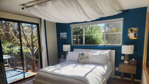 A bedroom with a bed, two bedside tables with lamps, a window with a view of trees, and a sliding glass door leading to an outdoor area.
