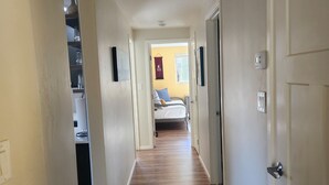 A narrow hallway with wooden flooring leading to a bedroom with a bed and a window.