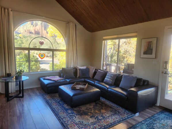 Una sala de estar moderna con un sofá modular, una mesa de centro redonda de vidrio y un amplio ventanal con vistas a árboles y un paisaje montañoso.