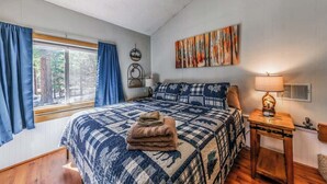 A bedroom with a large bed, a window with curtains, a wall-mounted lamp, and a painting.