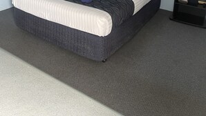 Ein ordentlich bezogenes Bett mit einem gemusterten Überwurf, zwei Kissen und einem gefalteten Handtuch. Ein schwarzer Nachttisch mit Schublade und Fernbedienung steht daneben. Die Wände sind hellblau und der Teppich ist grau.