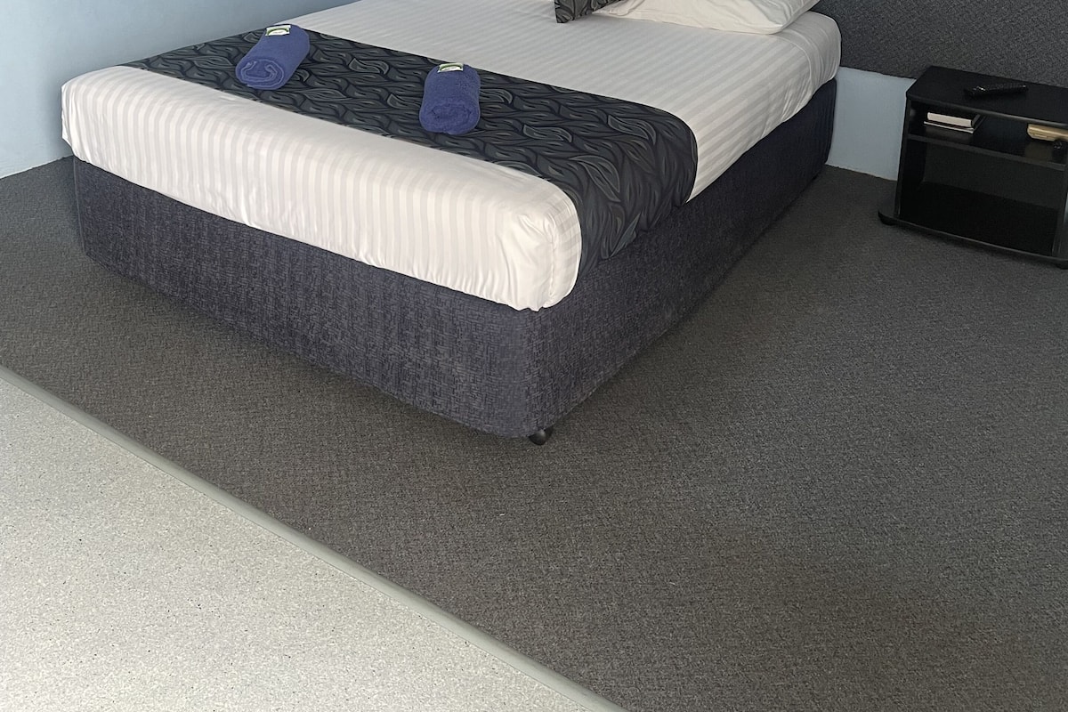 A neatly made bed with a patterned bedspread, two pillows, and a folded towel. There is a black nightstand with a drawer and a remote control. The room has light blue walls and a grey carpet.
