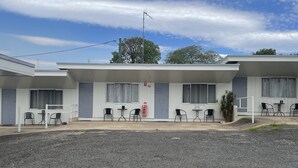 Ein Motel mit einer Reihe von Zimmern, jedes mit einem Fenster und einer Tür. Es gibt Tische und Stühle im Freien, und ein Hydrant ist sichtbar.
