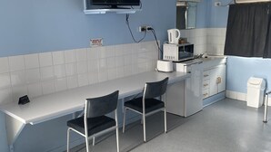 Eine kleine Küchenzeile mit Mikrowelle, Toaster und Wasserkocher. Ein Fernseher ist an der Wand befestigt, und ein Schild informiert über die Abreisebestimmungen. Zwei Stühle stehen an einem Tisch.