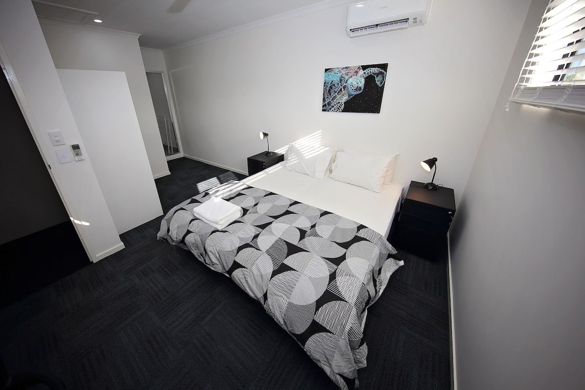 Una habitación de hotel moderna con una cama grande, una mesita de noche, aire acondicionado de pared y una obra de arte enmarcada sobre la cama.