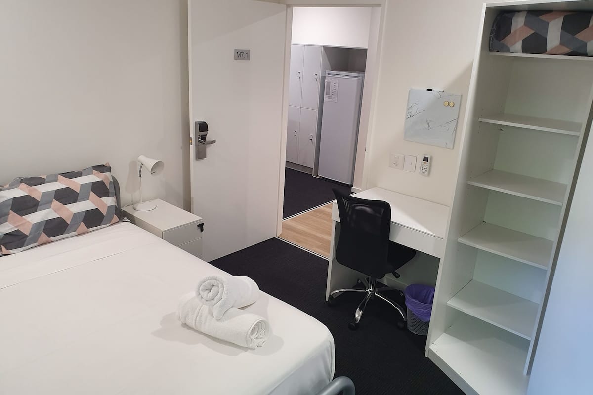 Habitación de hotel con cama, escritorio, silla, armario y baño visible a través de una puerta abierta.