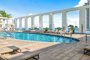 A pool area with lounge chairs and umbrellas, surrounded by a white structure with large windows.