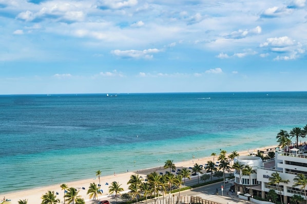 A coastal hotel with a beach view, palm trees, and a clear sky.