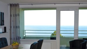A modern living room with a large window overlooking the sea, a dining area with a table and chairs, and a small balcony with a railing.