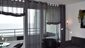 A modern living room with a black leather sofa, a black armchair, a glass coffee table, and sheer curtains over the windows.