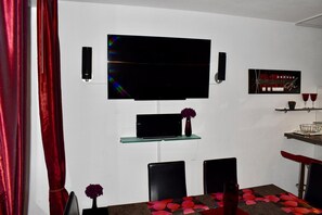 Un salon moderne avec un téléviseur à écran plat fixé au mur, une étagère en verre en dessous, et un coin repas avec une table mise pour deux personnes.