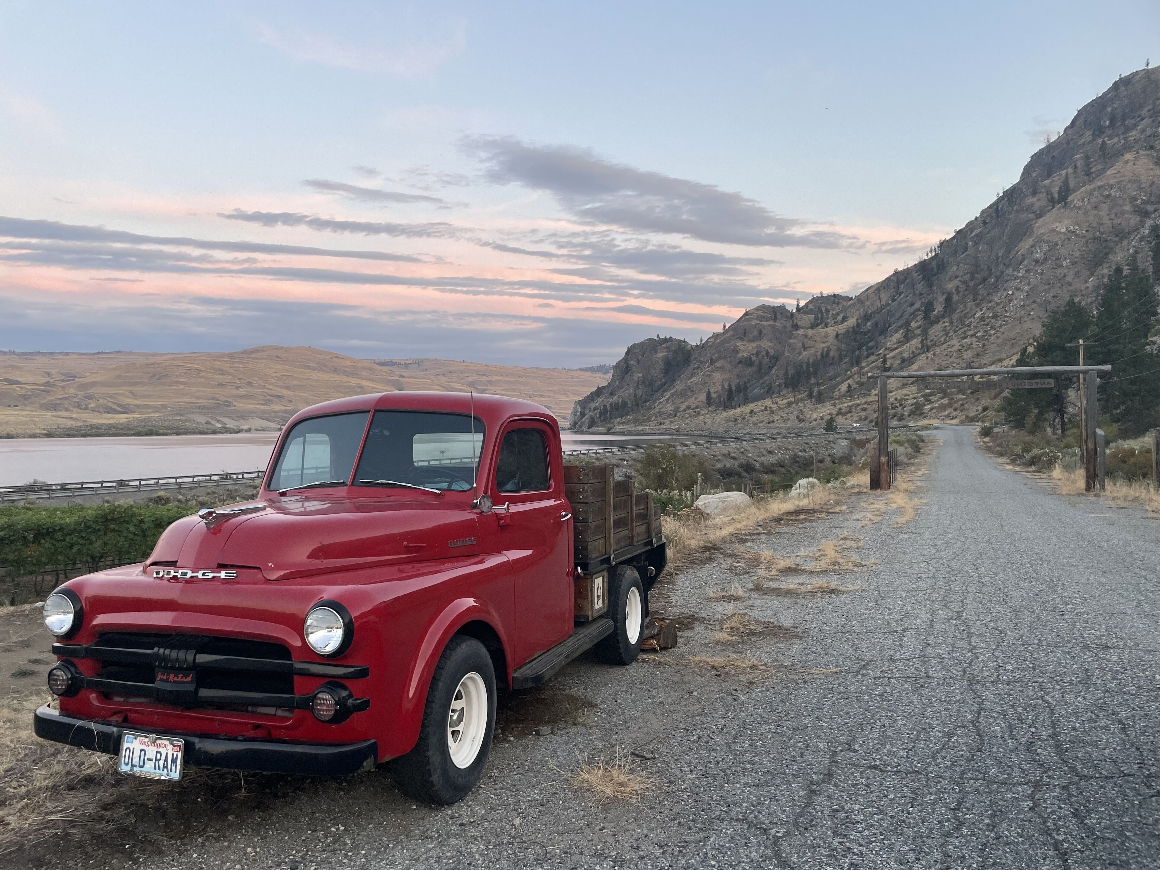 A red vintage Dodge pickup truck parked on a gravel road with a scenic view of hills and a body of water in the background.