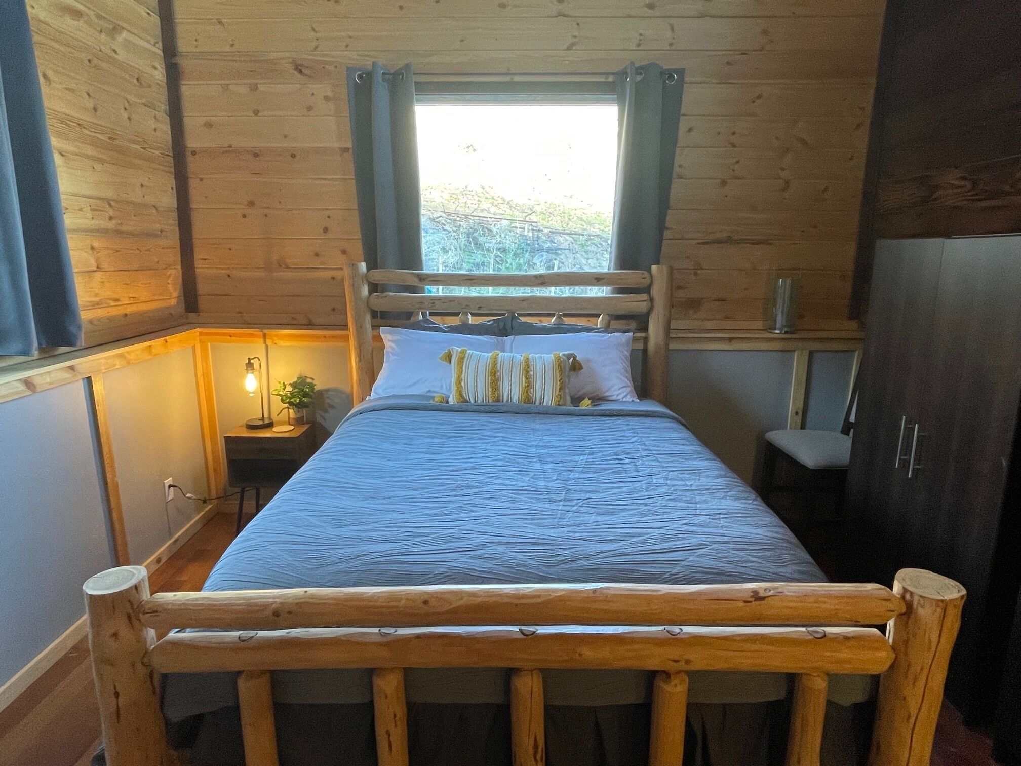 A wooden cabin bedroom with a bed, a nightstand, a lamp, and a window with curtains.