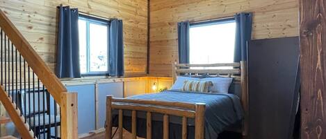 A wooden cabin room with a bed, a ceiling fan, and a window with curtains.
