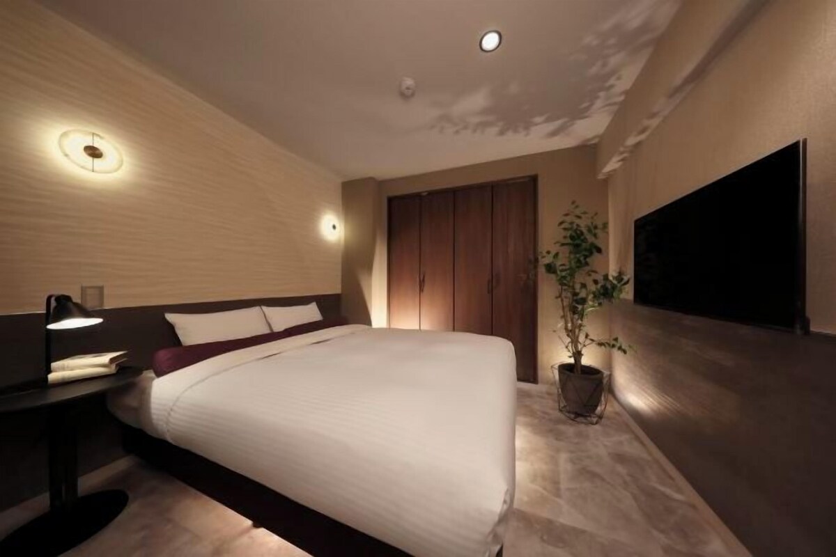 A modern hotel room with a large bed, a flat-screen TV mounted on the wall, a nightstand with a lamp, and a potted plant.