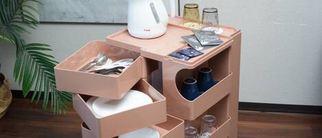 Un coin cuisine comprenant une bouilloire blanche, un plateau avec des verres et un paquet, ainsi qu’une étagère avec des tasses et des assiettes.