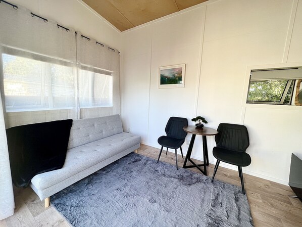 Ett modernt rum med en grå soffa, två svarta stolar, ett litet runt bord med en bonsai och ett stort fönster med tunna, genomskinliga gardiner.