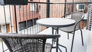 Een balkon met een ronde tafel en twee rieten stoelen, een zwarre metalen hekwerk en uitzicht op een bakstenen gebouw.