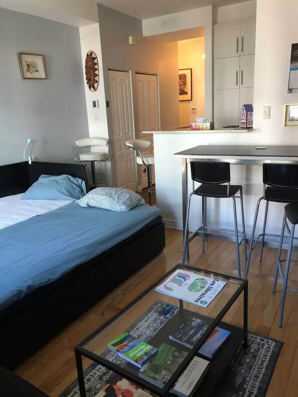 Una habitación compacta con cama, una pequeña mesa de centro con libros, una kitchenette y un taburete de bar.
