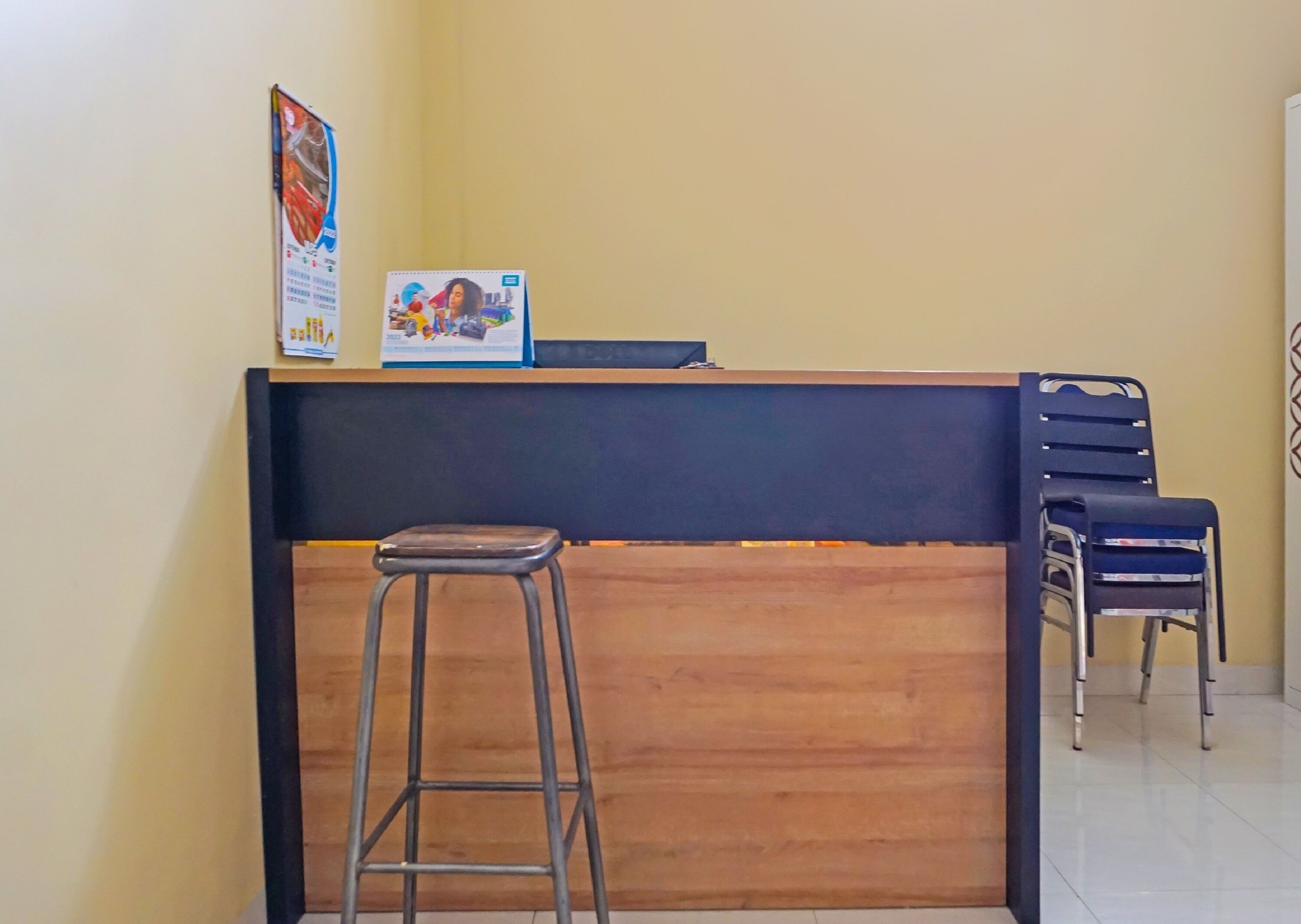 A reception desk with a wooden counter and a metal stool in front. There are two chairs to the right of the desk.