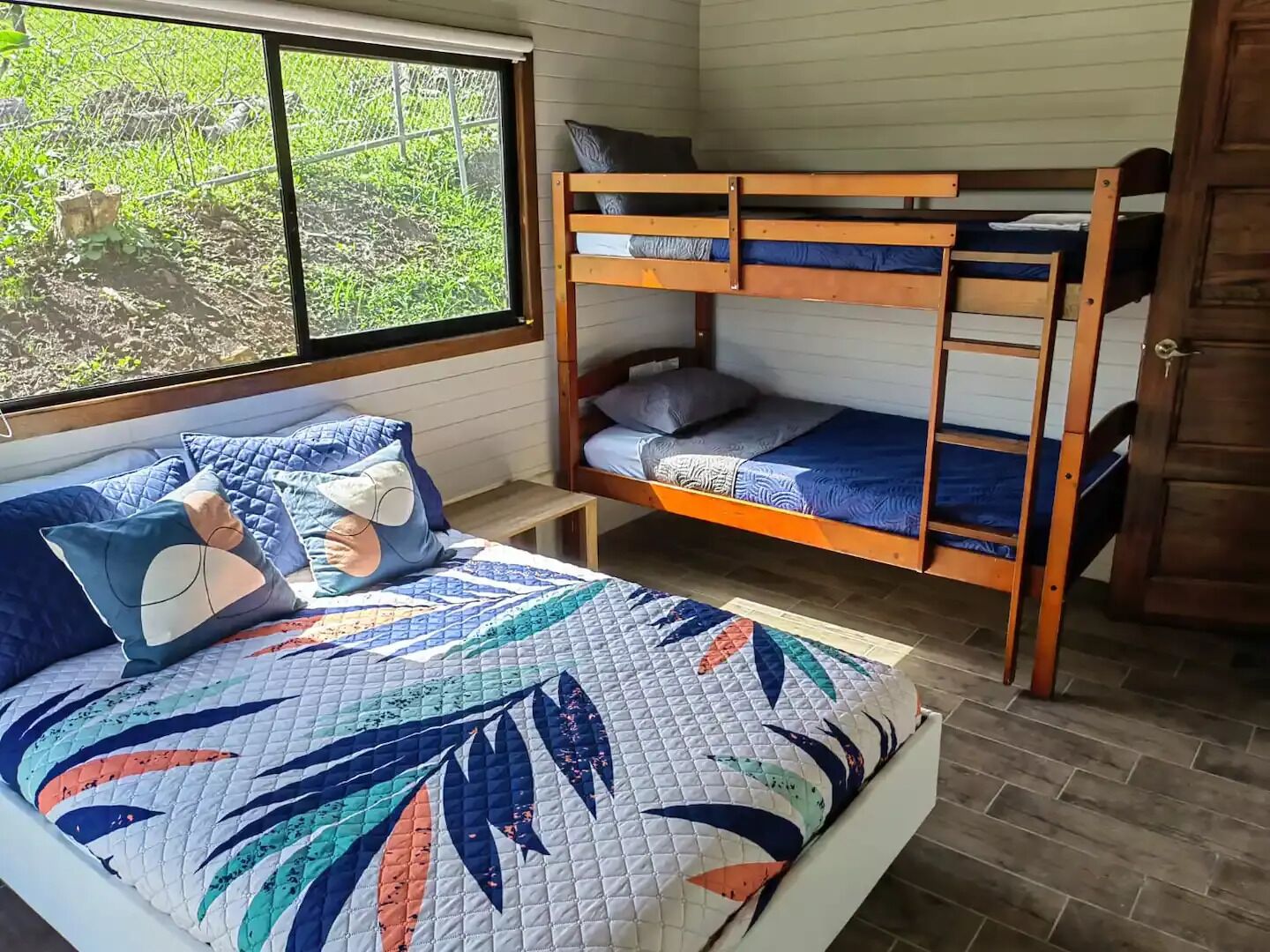 Un dormitorio con una litera, una cama individual con colcha colorida y un ventanal con vistas a la vegetación.
