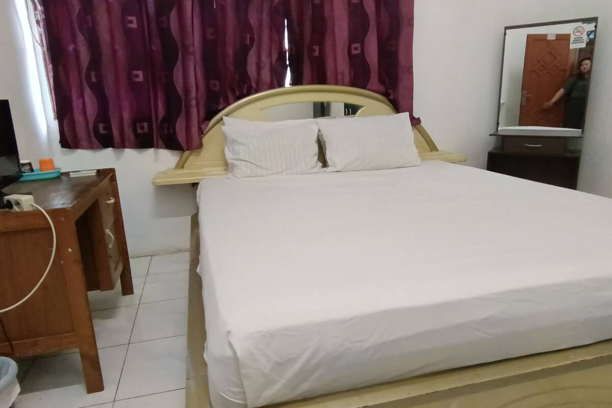 A neatly made bed with white linens, a wooden desk with a computer and a telephone, and a mirror on the wall.