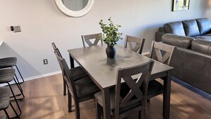 Un comedor con una mesa y sillas de madera, una planta en maceta, un espejo redondo en la pared y un sofá gris al fondo.