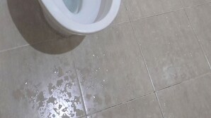 Un inodoro blanco con la tapa cerrada, el botón de descarga a la vista y una tubería visible.