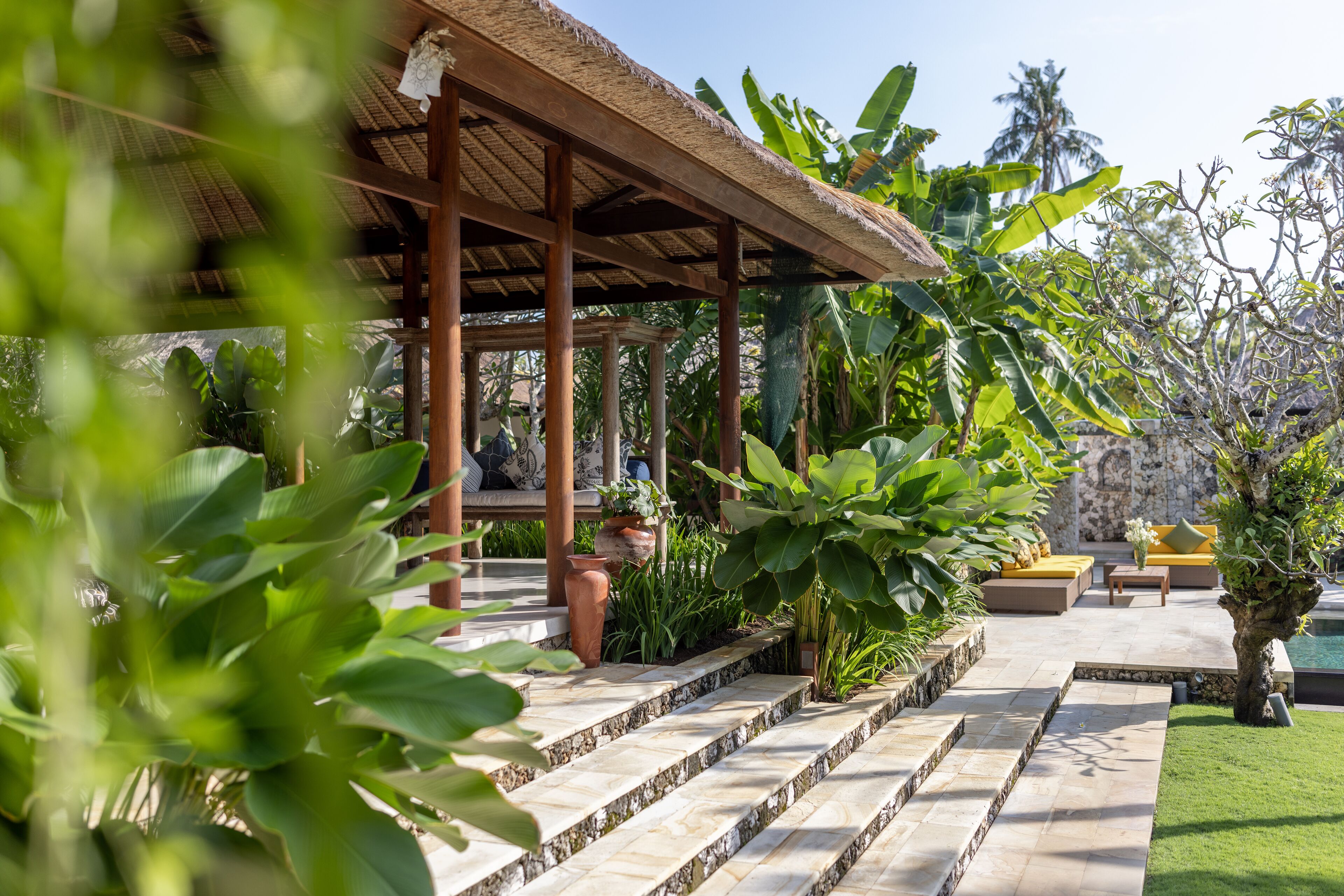 A covered outdoor seating area with stone steps leading to a seating arrangement, surrounded by lush greenery and a pool.