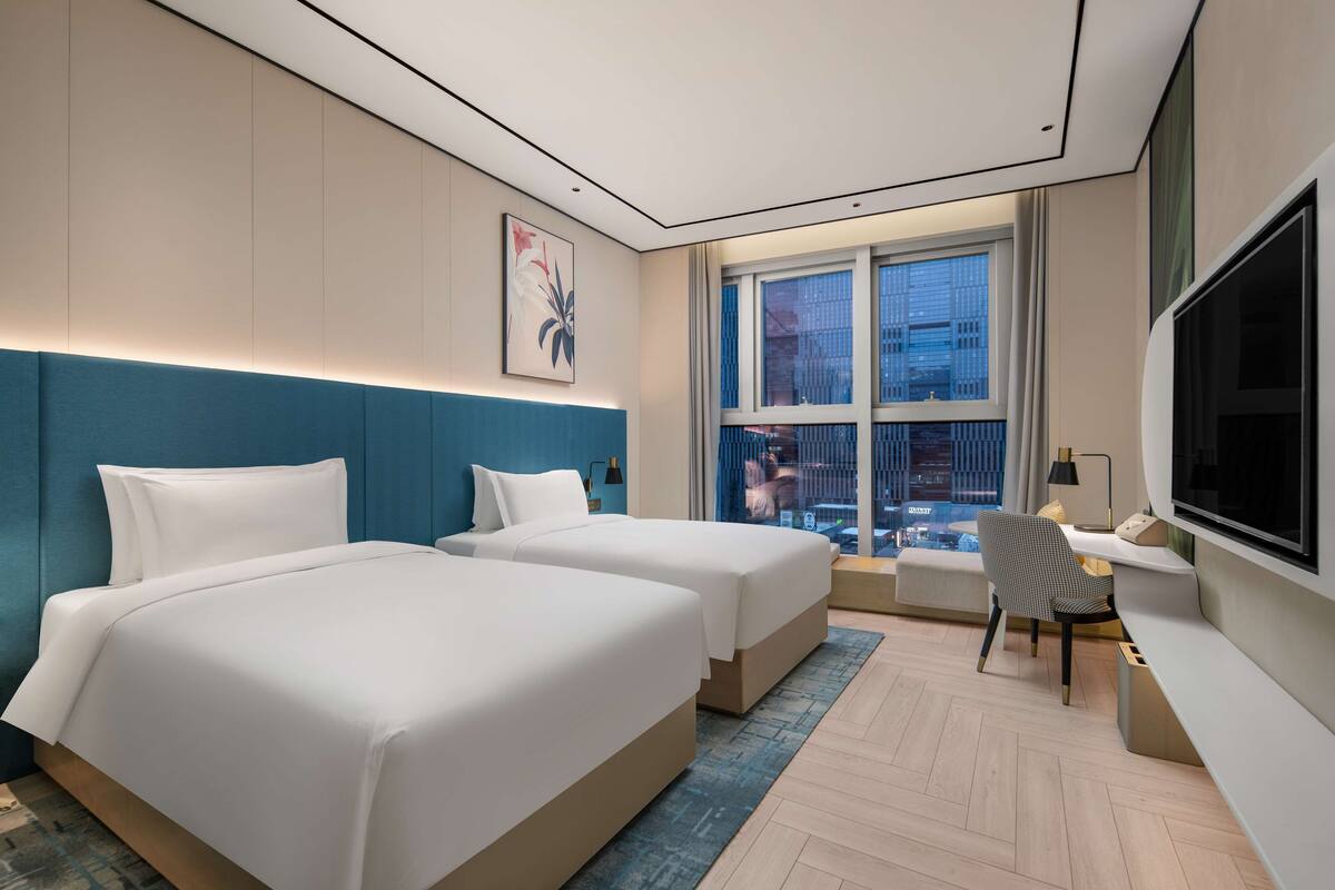 Une chambre d’hôtel avec deux lits, un bureau, une chaise, une télévision et une grande fenêtre donnant sur la ville.