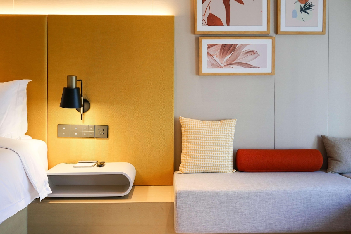 A hotel room with a bed, bedside table, lamp, wall-mounted power outlets, and artwork.