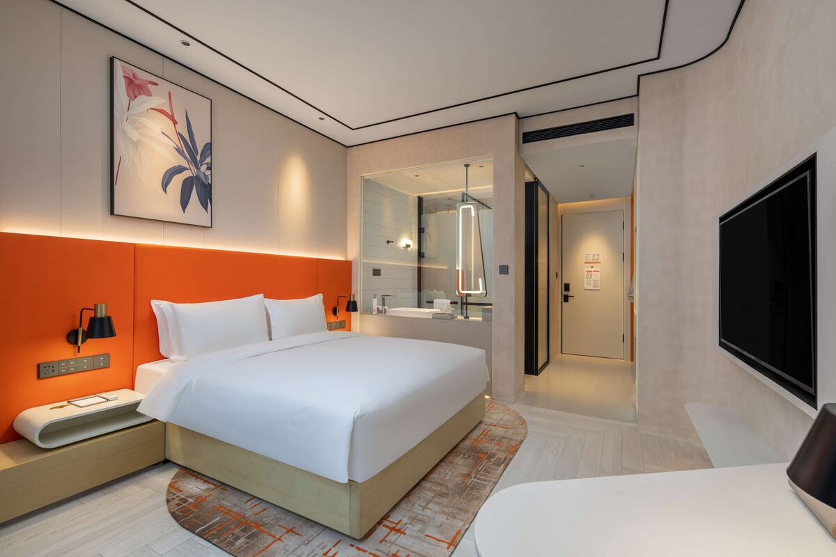 Une chambre d’hôtel moderne dotée d’un grand lit, d’une table de chevet avec une lampe, d’un téléviseur à écran plat et d’une salle de bain visible à travers une paroi en verre.