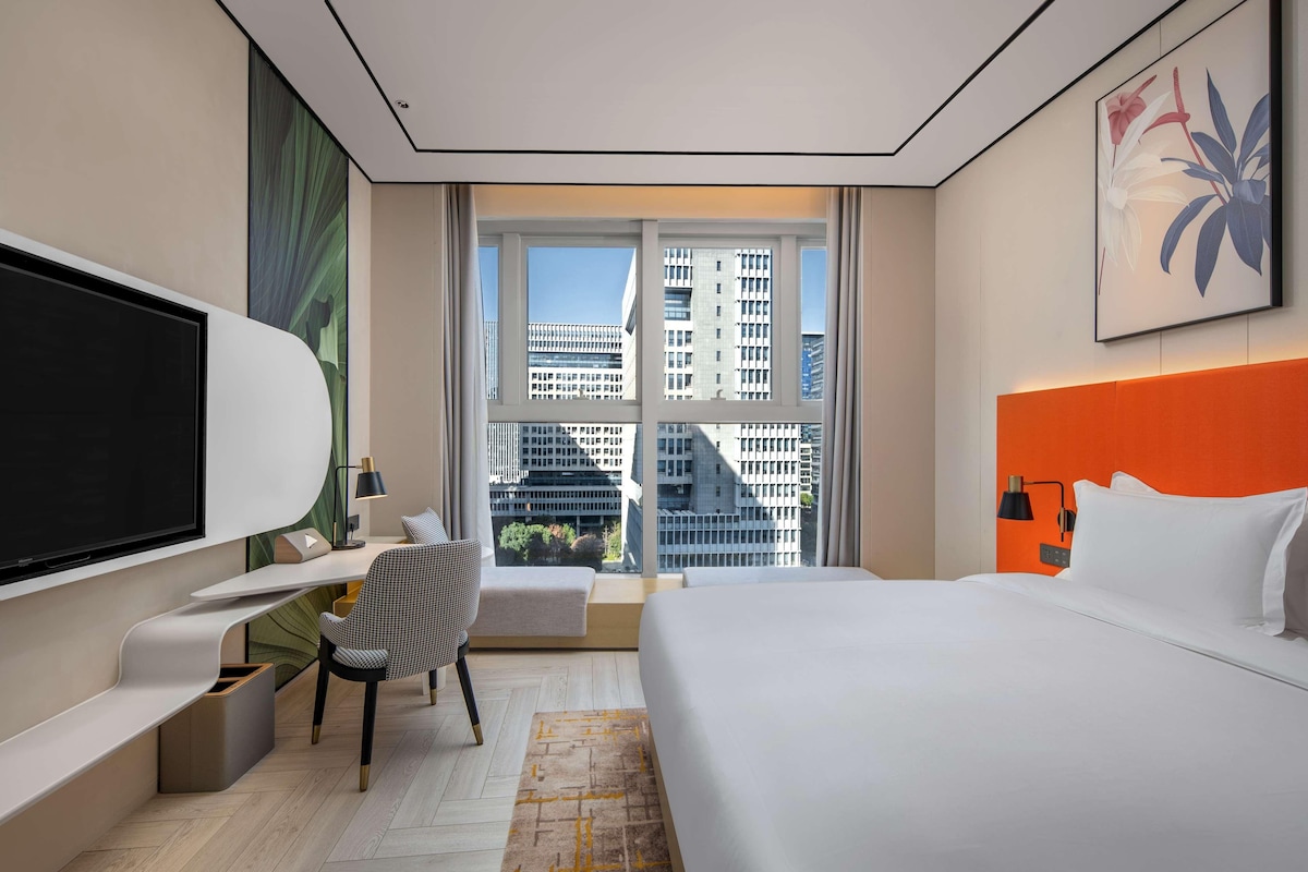 A modern hotel room with a large bed, a desk with a chair, a TV, and a window with a city view.