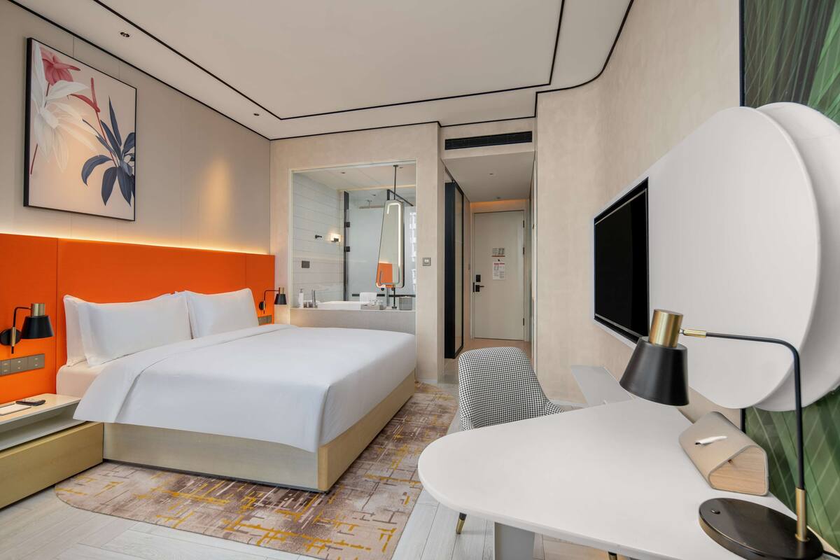 Une chambre d’hôtel moderne, dotée d’un grand lit, d’un bureau, d’une télévision et d’une salle de bain visible à travers une porte ouverte.
