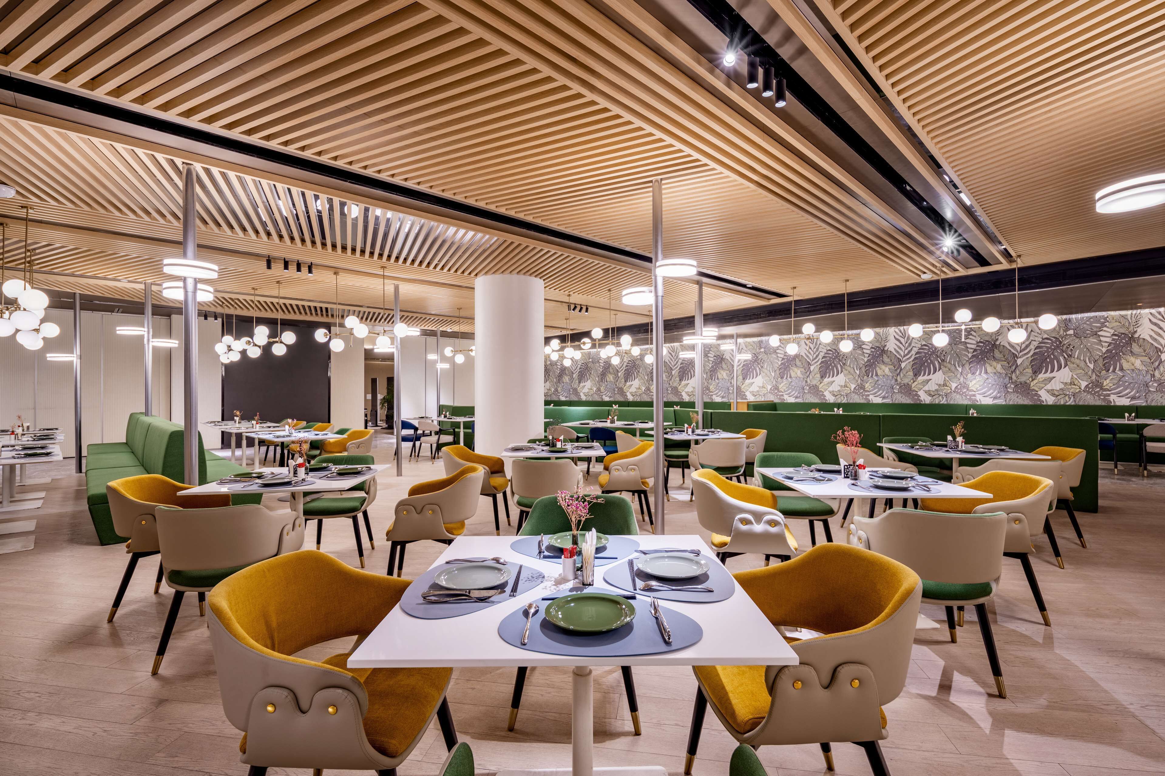 Un restaurant moderne avec des panneaux de plafond en bois, des chaises vertes et jaunes, et des tables dressées pour le service.