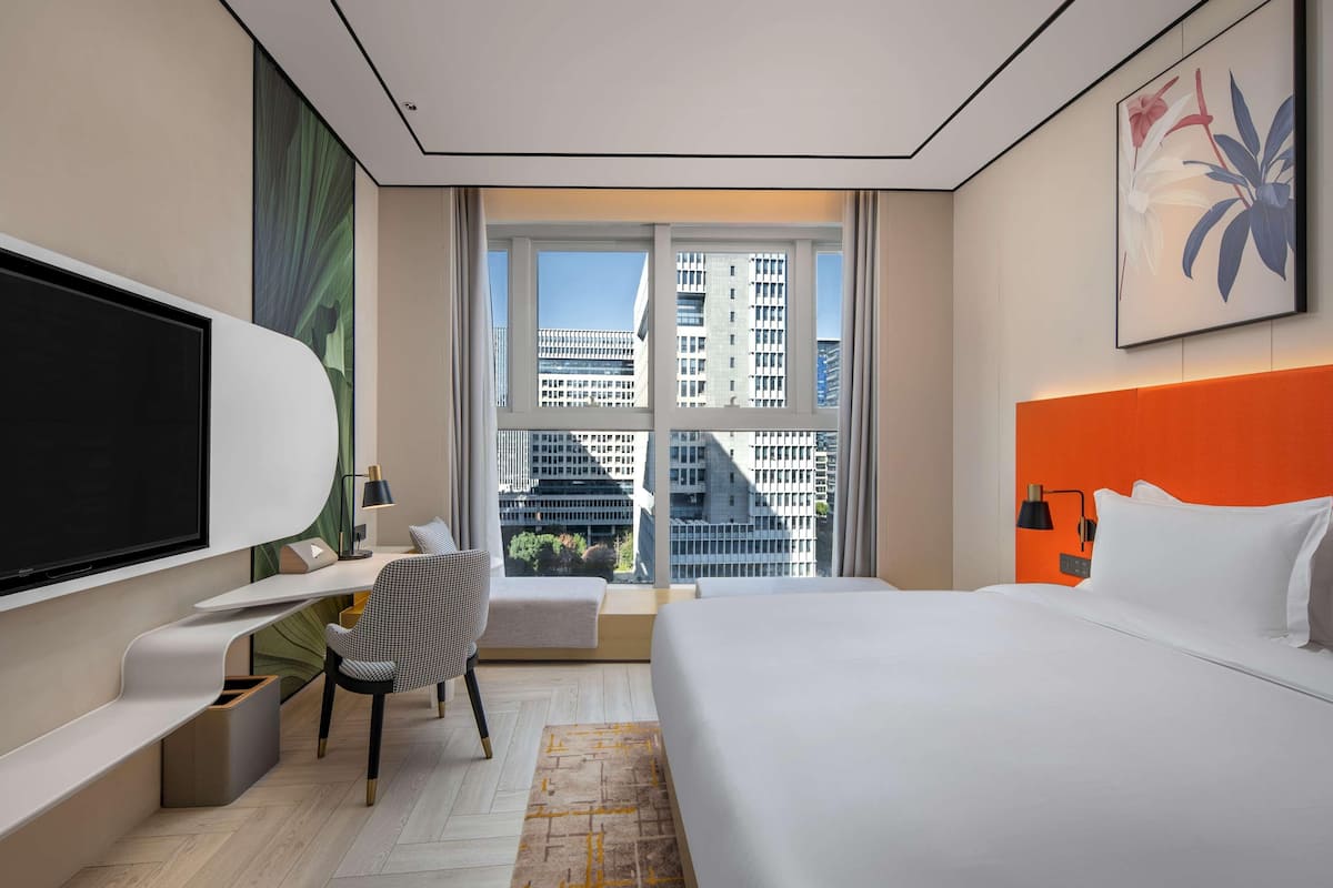 Une chambre d’hôtel moderne dotée d’un grand lit, d’un bureau avec une chaise, d’une télévision et d’une fenêtre donnant sur la ville.